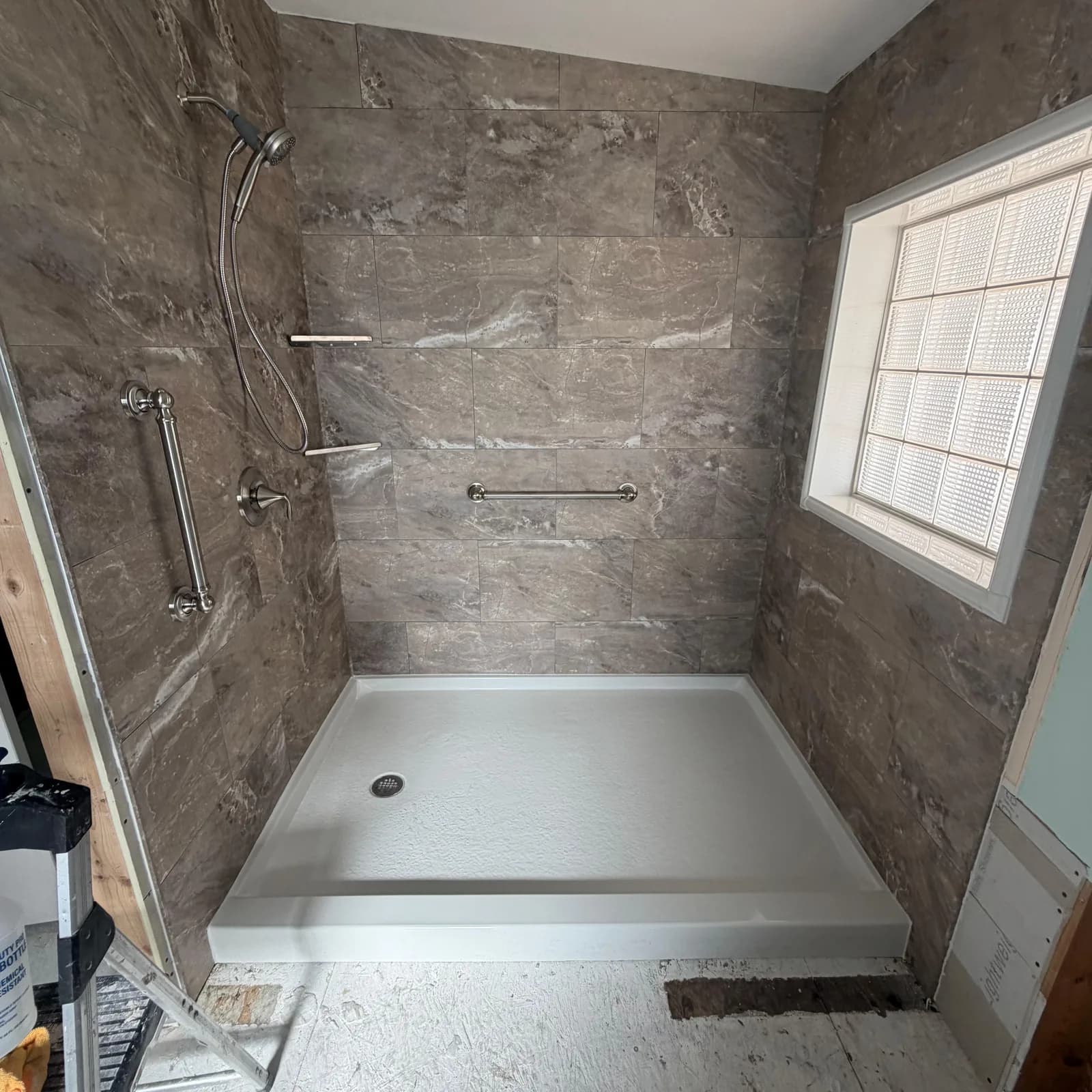 An accessible walk-in shower mid-build, with grab bars set, stone-look tile installed, and the job site still in progress