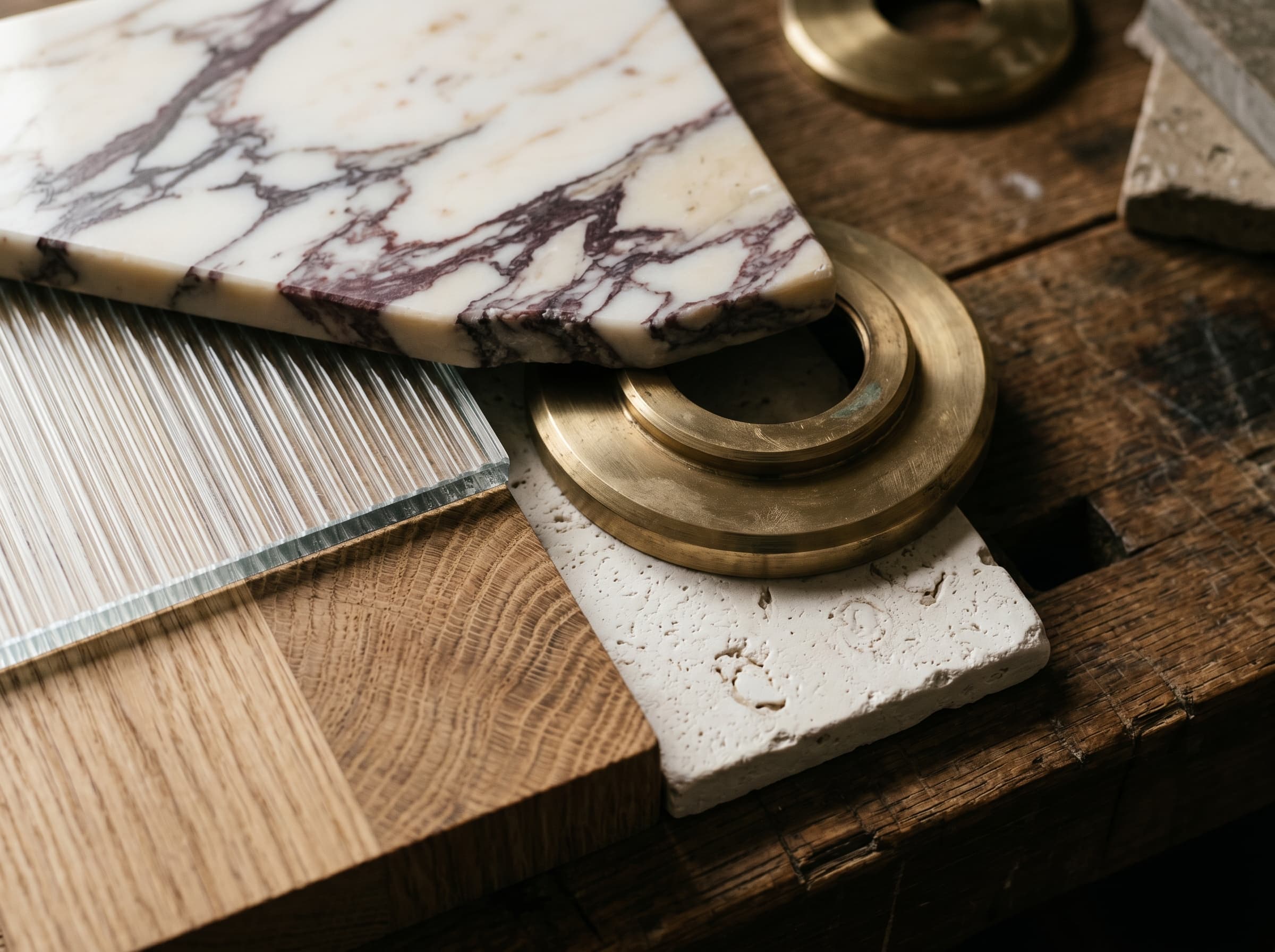 A craft moodboard of Calacatta Viola marble, fluted glass, champagne brass, oak, and travertine on an oak workbench