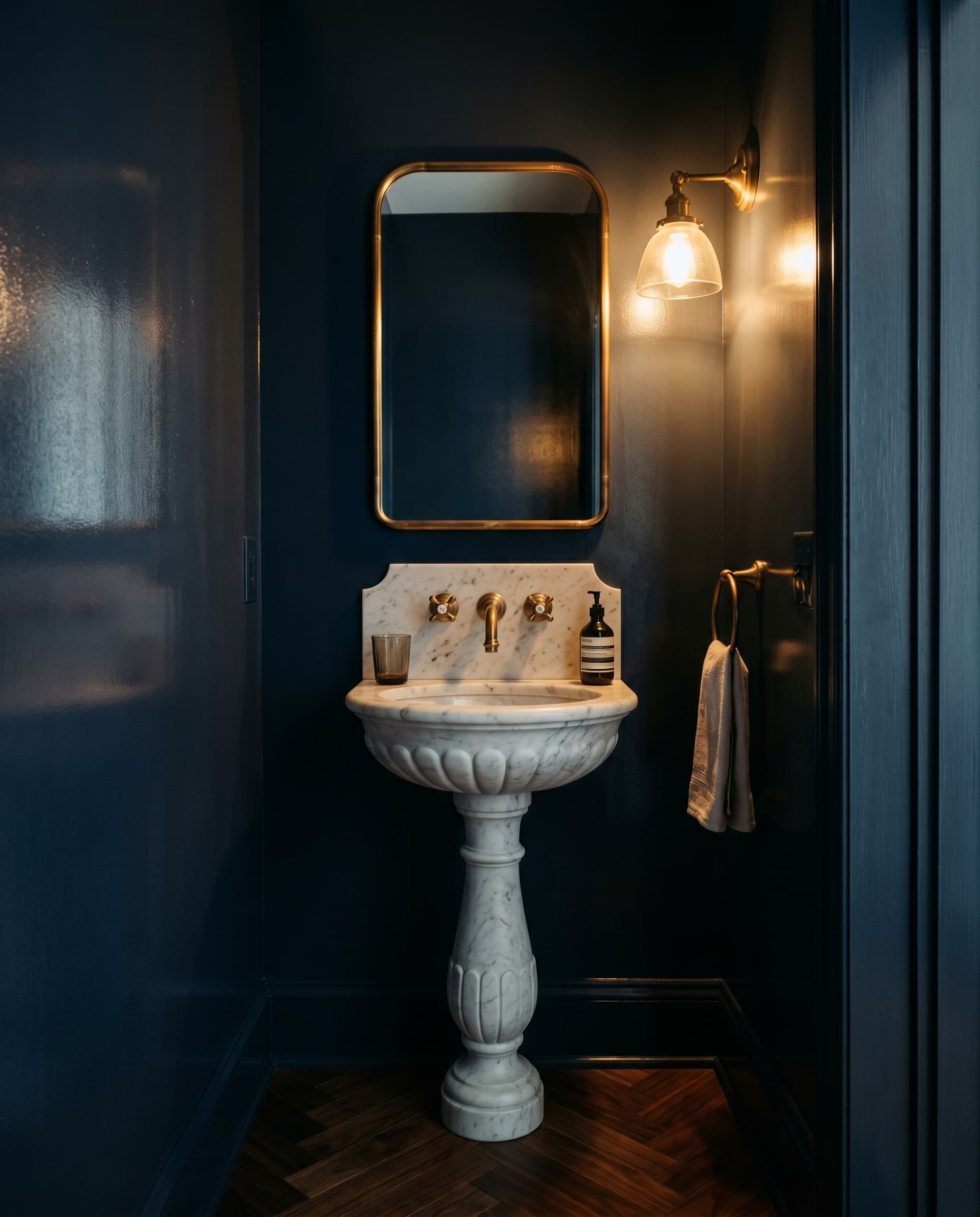 Deep navy powder room with marble pedestal sink, brass cross-handle faucet, and herringbone walnut floor